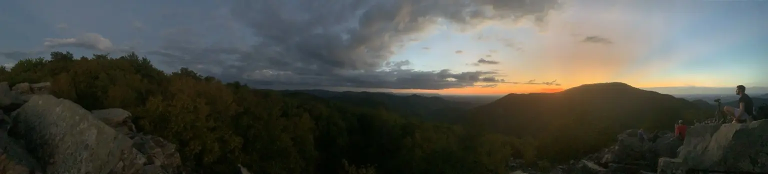 Panoramic sunset over a forested mountain ridge with hikers resting on the rocks