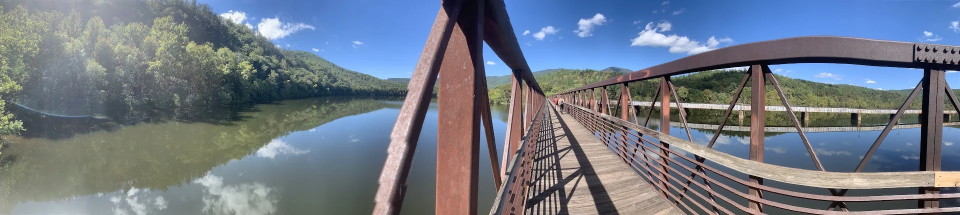 Panoramic view from a metal footbridge across a calm river surrounded by wooded hills